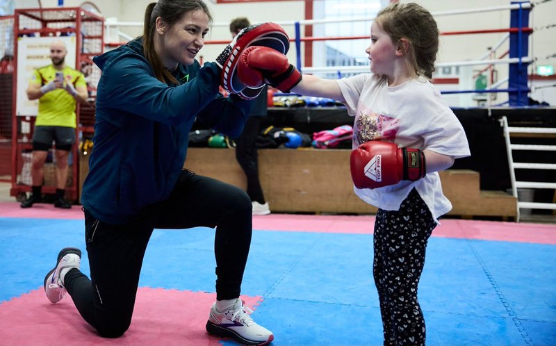 A coach kneeling to hold the pads for a small girl in boxing gloves